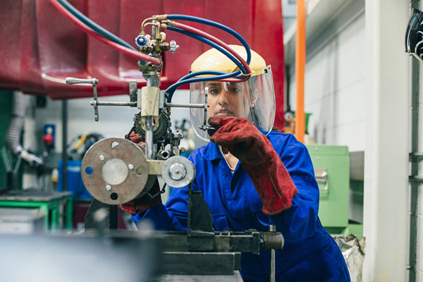 A young woman is using engineering equipment in a workshop. She is wearing a protective mask and gloves as well as blue coveralls.